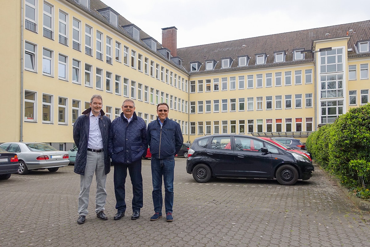 Drei Männer stehen vor einem gelben Bürogebäude mit mehreren Fenstern und geparkten Autos auf dem Parkplatz